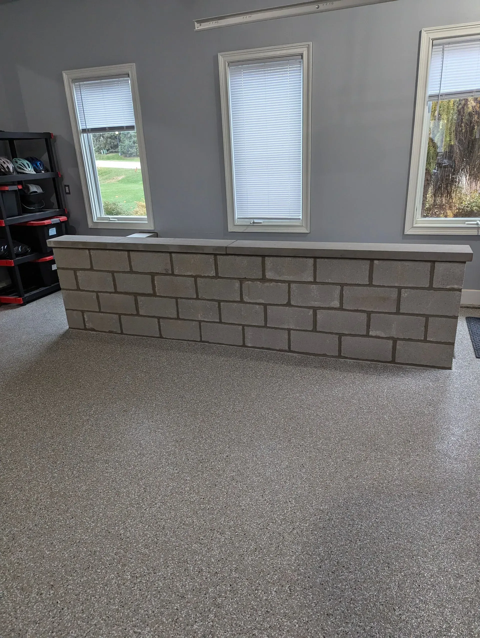 A gray cinder block wall stands in a room with speckled flooring and three windows with white blinds on a gray wall.