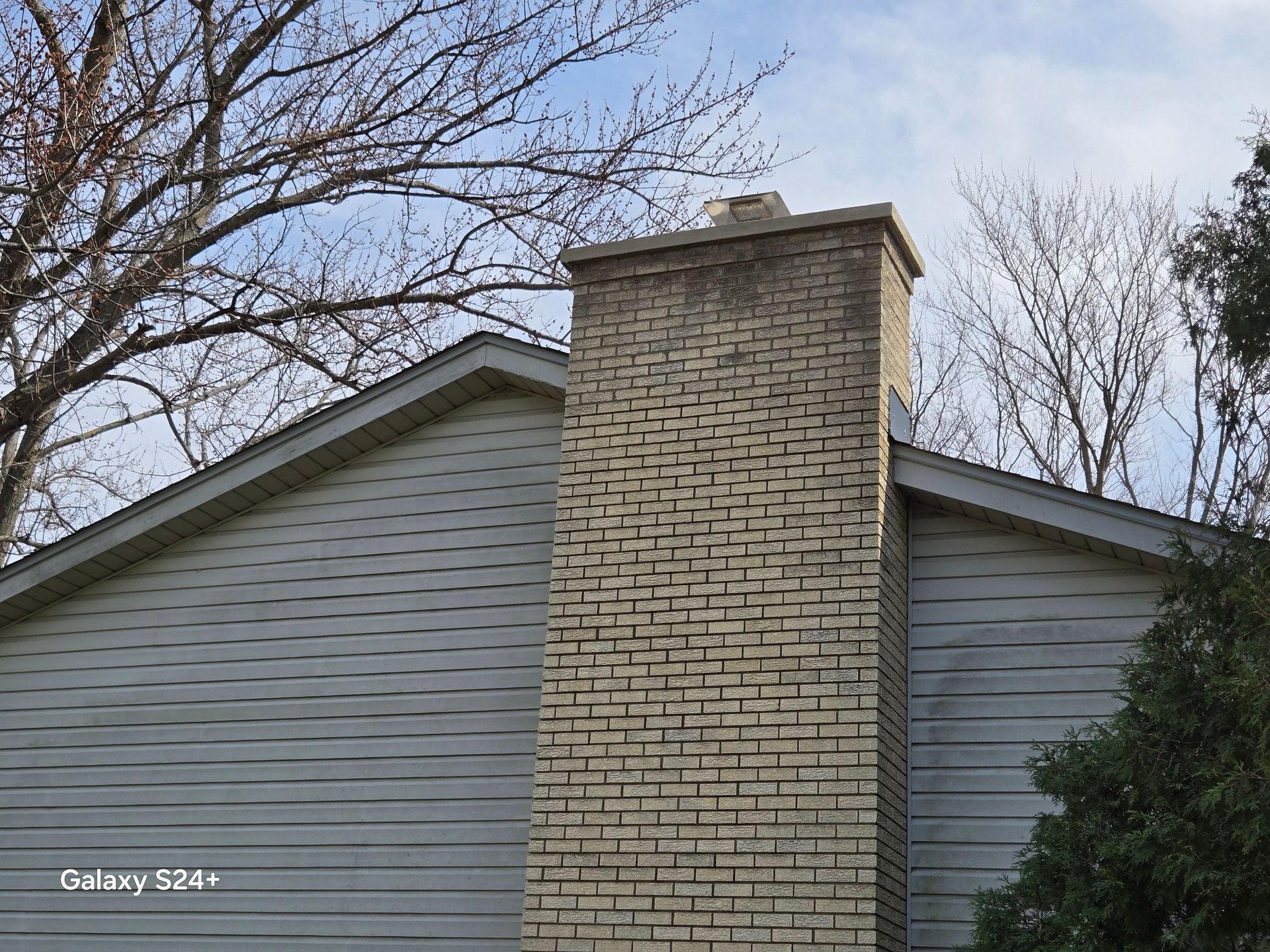 A light-colored brick chimney rises between two beige siding walls against a backdrop of trees and a blue sky.