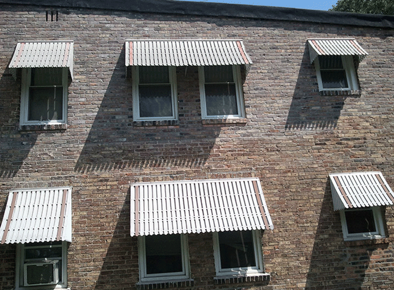 A brick wall with five windows, each covered by a white metal awning, arranged in two rows.