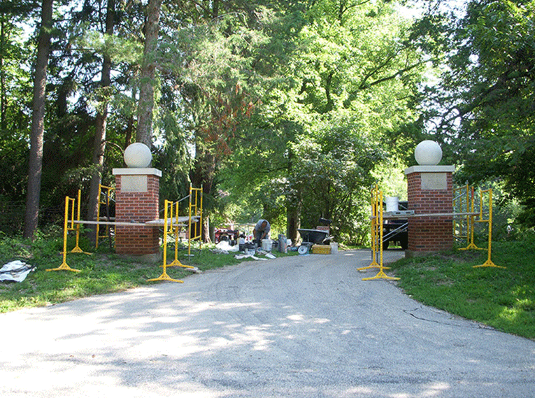 Brick pillars with spheres on top flank a driveway entrance, surrounded by yellow construction scaffolding and greenery.