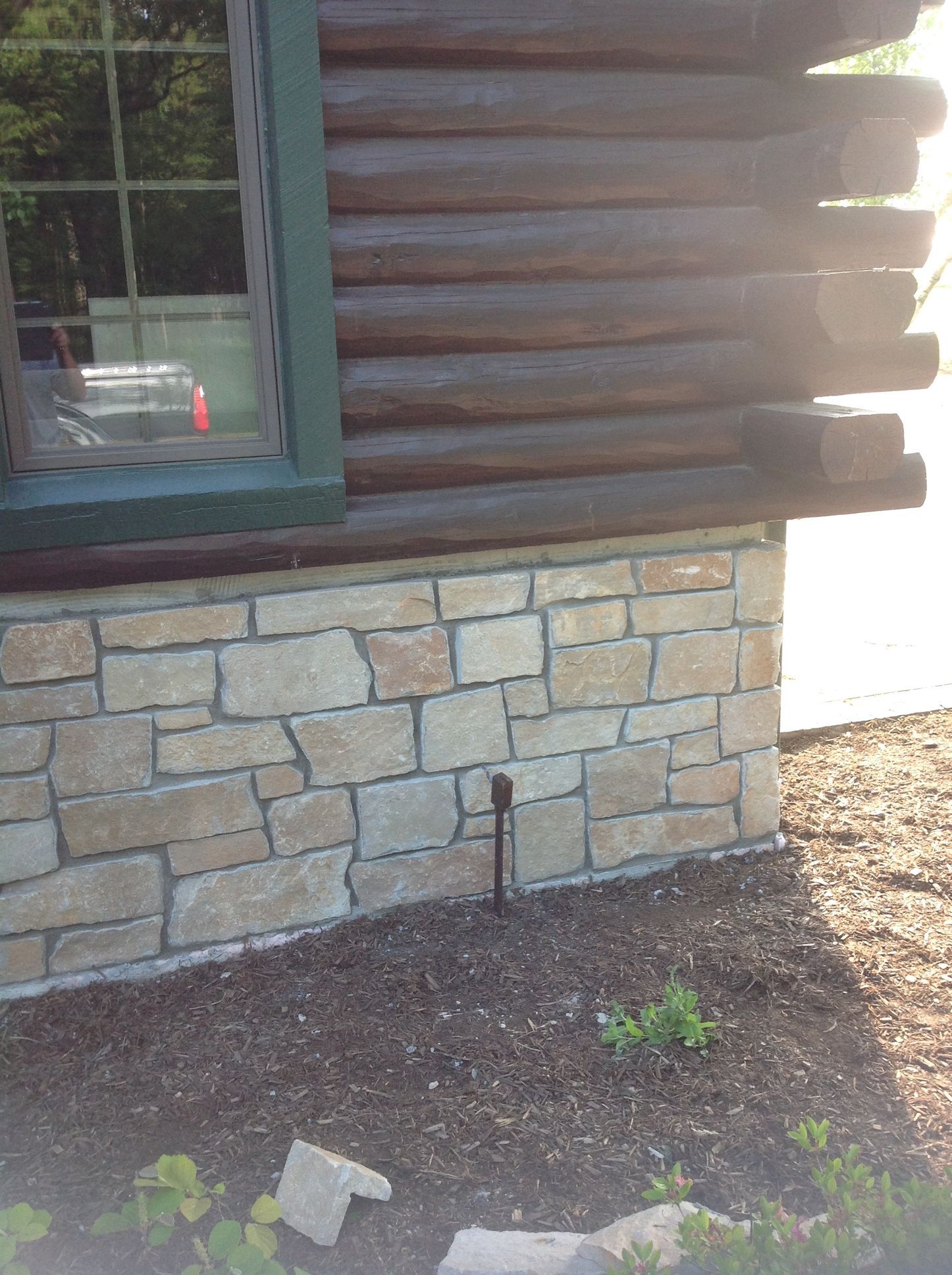 A corner view of a log cabin exterior featuring a stone foundation and a window with green trim.