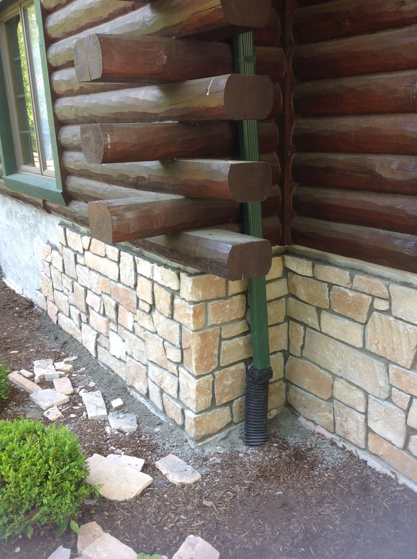 A log cabin corner with stone foundation and a green downspout against the wall, with scattered stone debris on the ground.