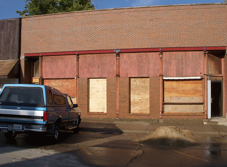 A blue pickup truck parked in front of a brick building with plywood-covered windows and an open doorway.