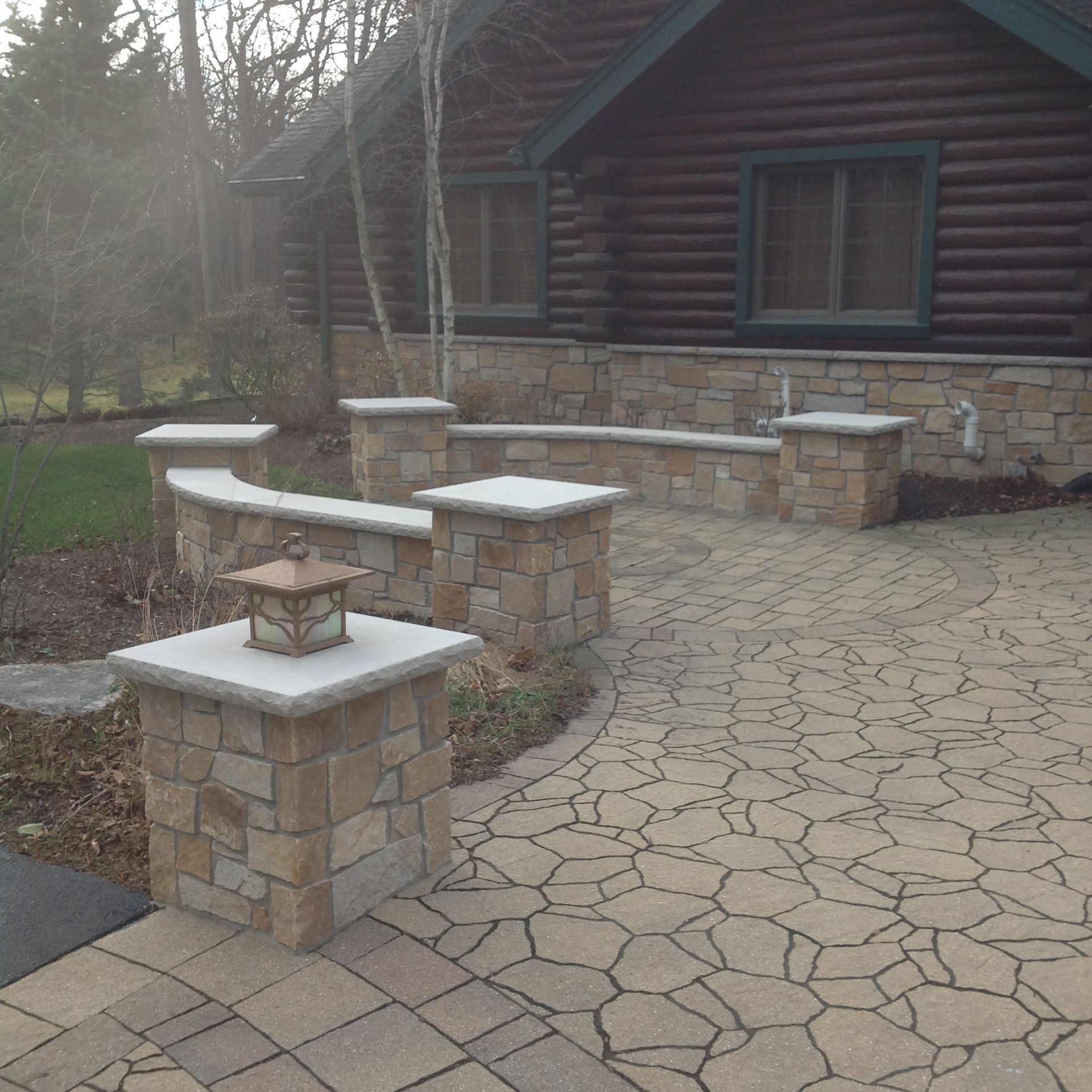 A stone patio with low seating walls and square stone pillars, situated in front of a rustic log cabin.