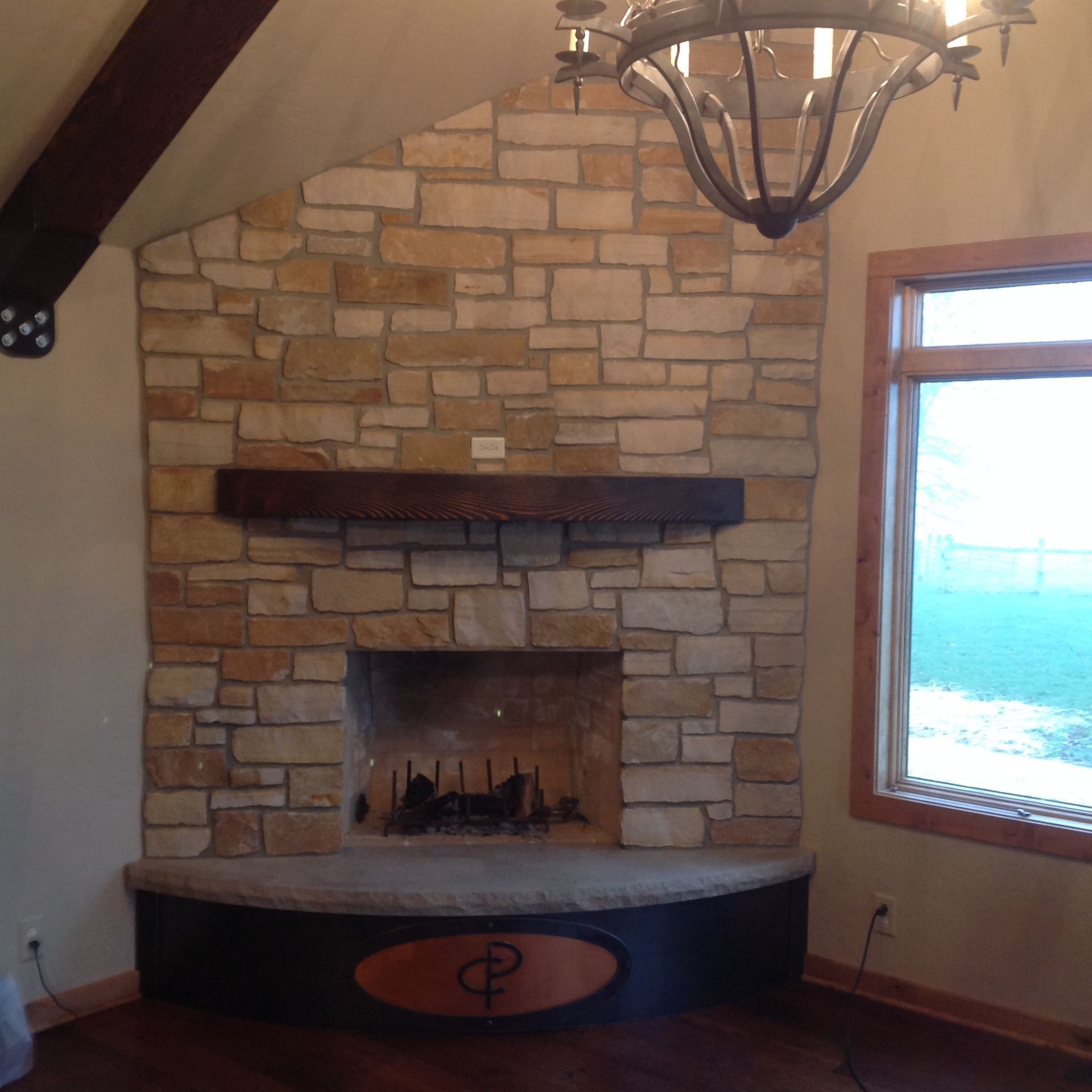 A rustic stone fireplace with a dark wood mantel and a curved hearth featuring an oval wooden logo insert.