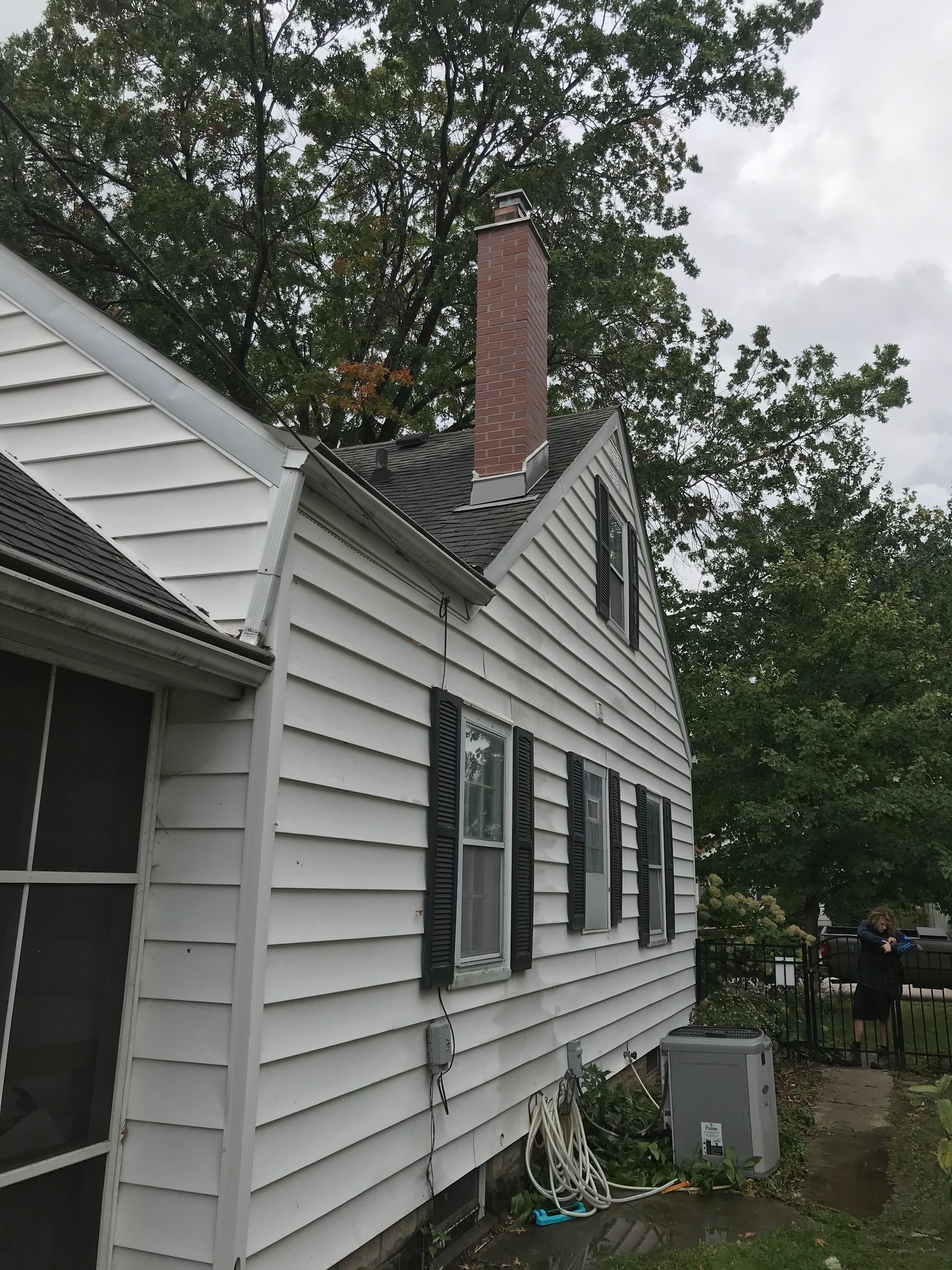 Side view of a white, two-story house with black shutters, a brick chimney, and a tree in the background.