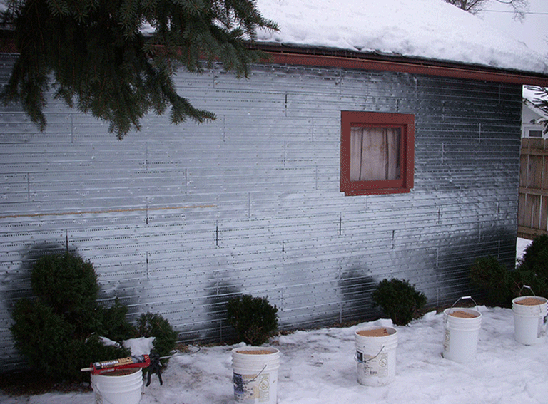 A garage exterior covered in silver reflective insulation, with several white buckets placed along a snow-covered yard.