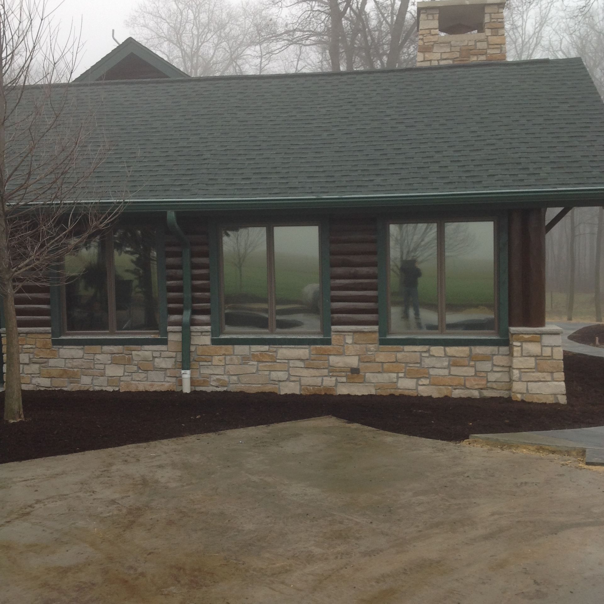 A log cabin with a stone base, green roof, and three large windows, viewed on a foggy day.
