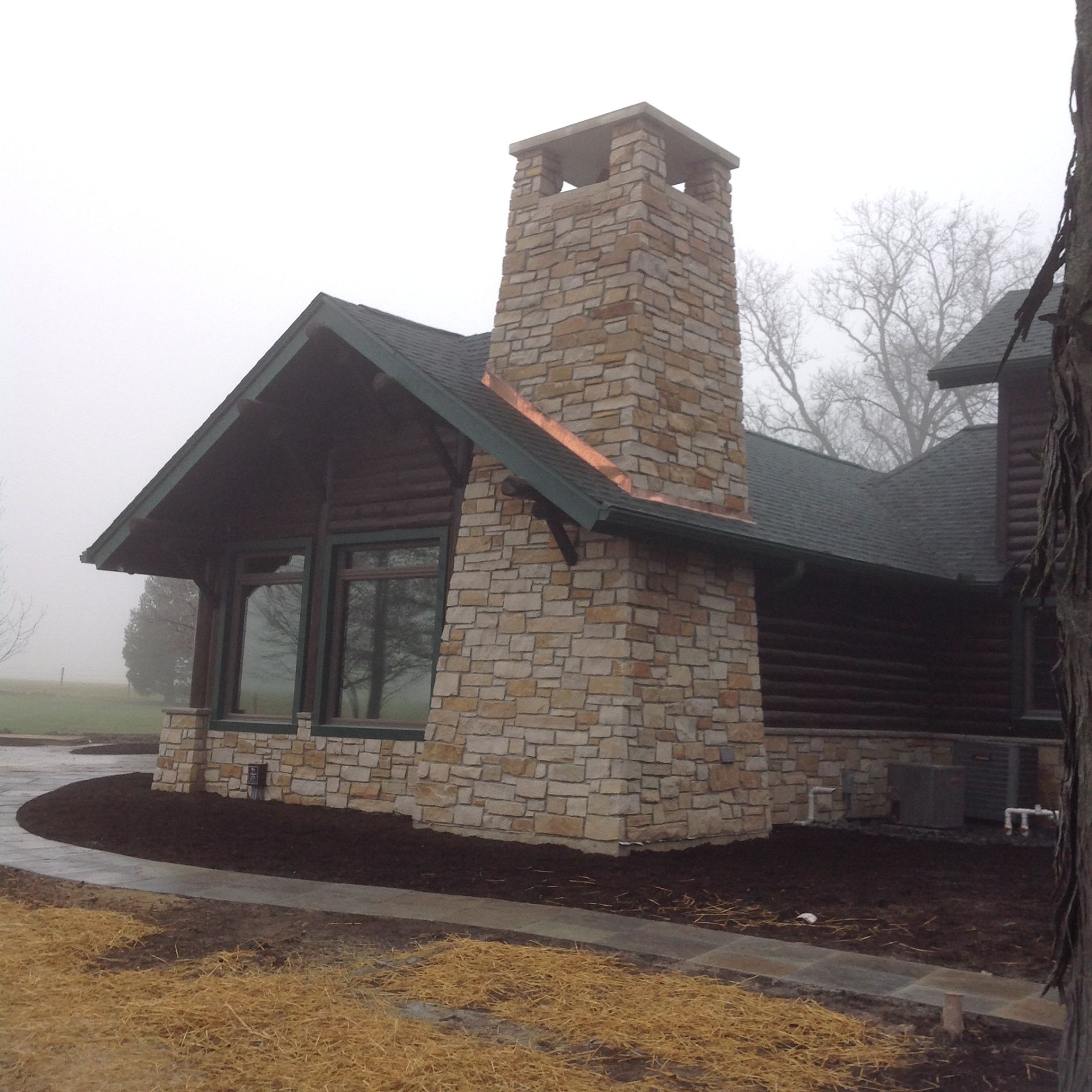 A cabin with a large stone chimney exterior, featuring dark log siding and a shingled roof on a foggy, overcast day.