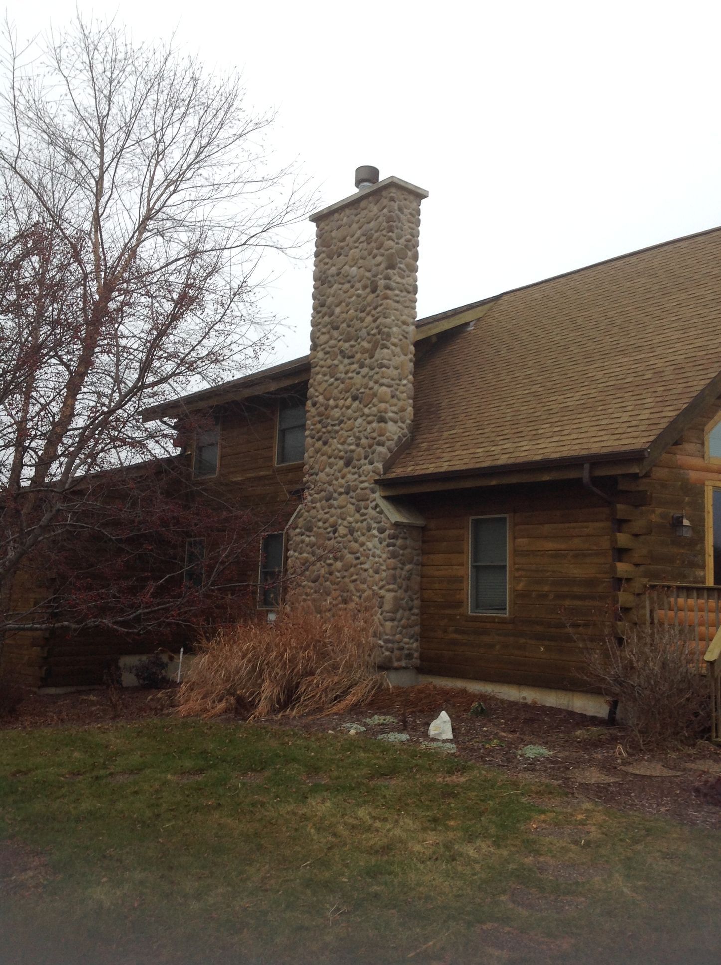 A log cabin exterior with a large stone chimney extending above the brown shingled roof, set against a cloudy sky.