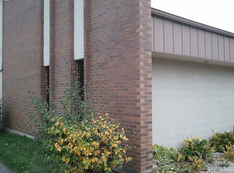 Exterior view of a brick building with white vertical accents, a brown metal roof section, and shrubs in the foreground.