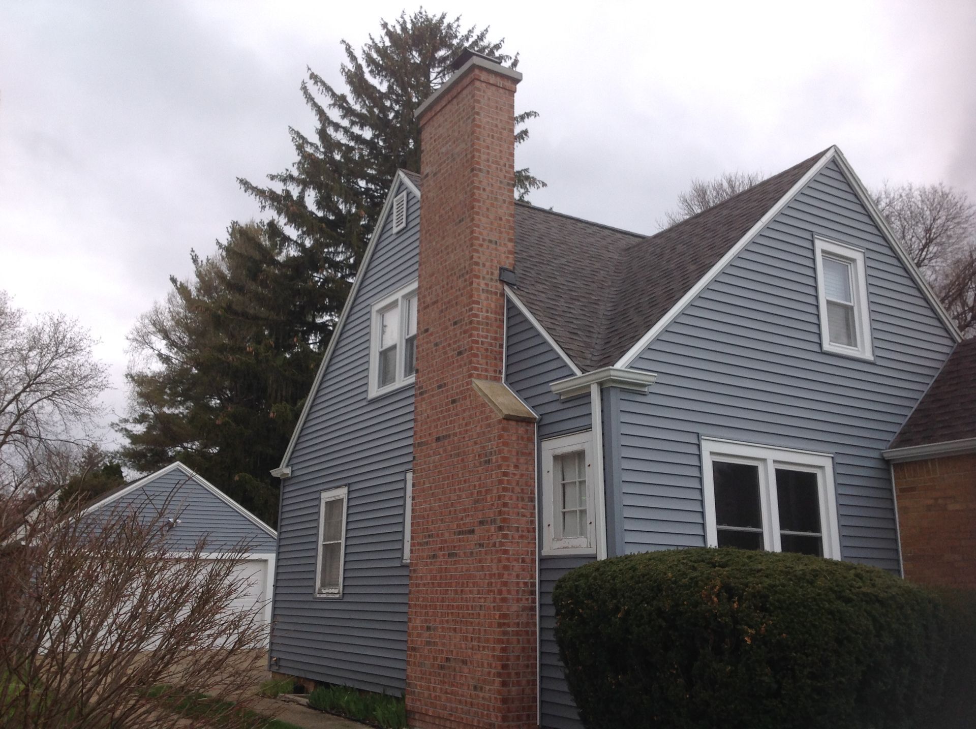 A blue-gray house with a brick chimney and an adjacent detached garage under a cloudy sky.