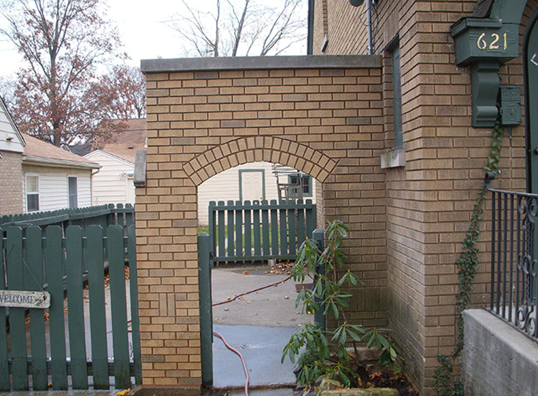 A tan brick building with an arched gate leading to a backyard, with a green wooden fence and a small evergreen plant.