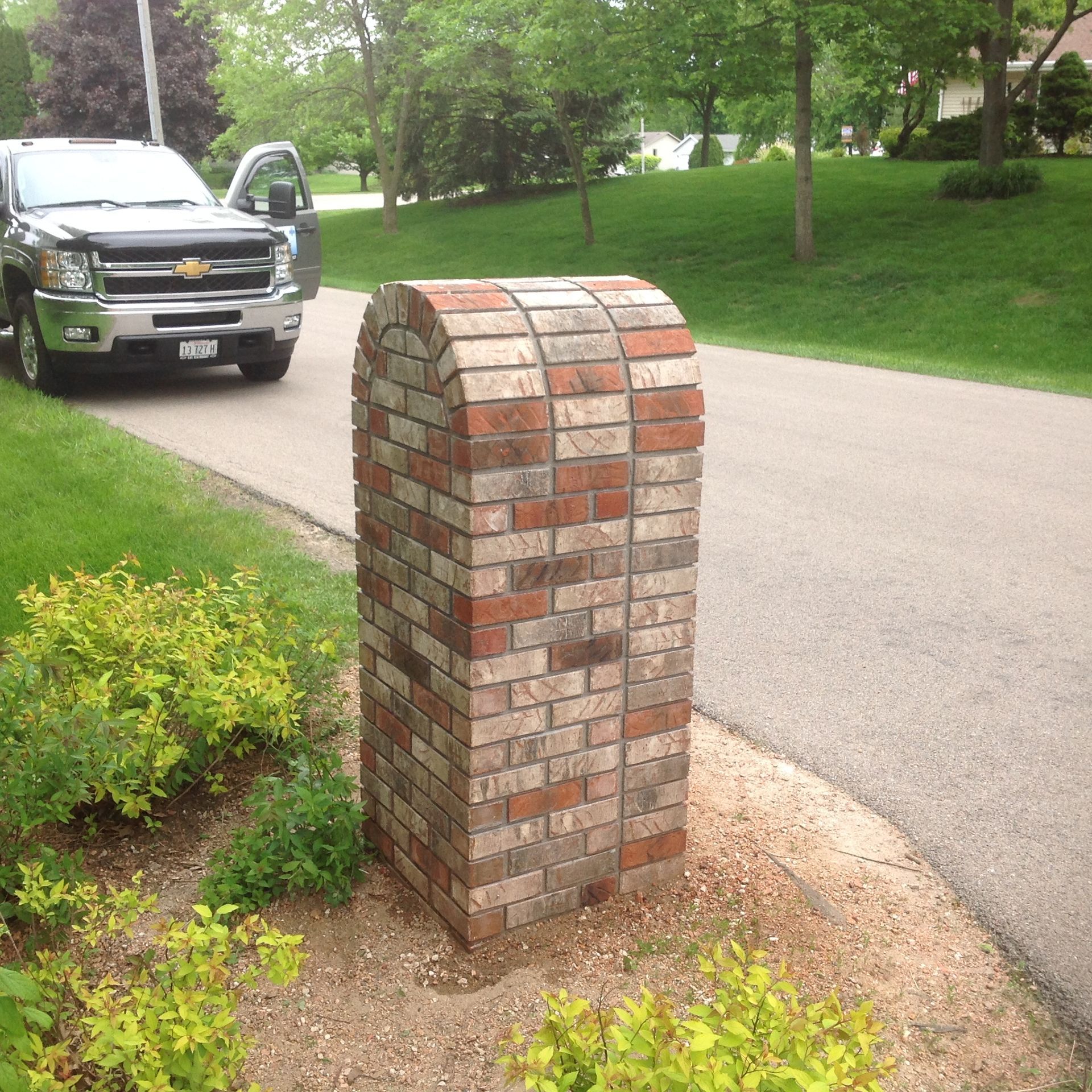 A multi-colored brick mailbox structure sits near a paved driveway with a truck parked in the background.