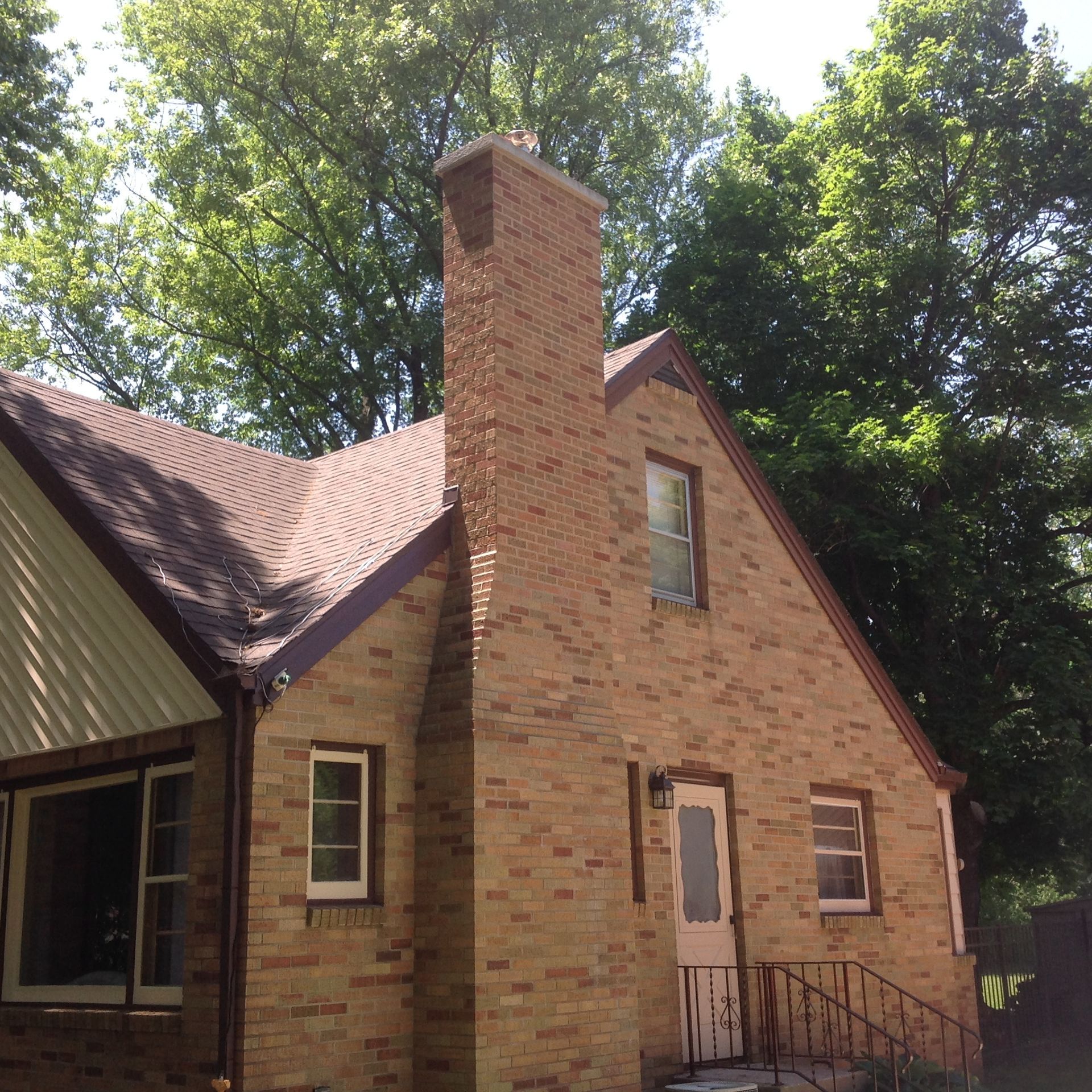 A brick house with a prominent chimney, dark shingled roof, and green trees in the background on a sunny day.