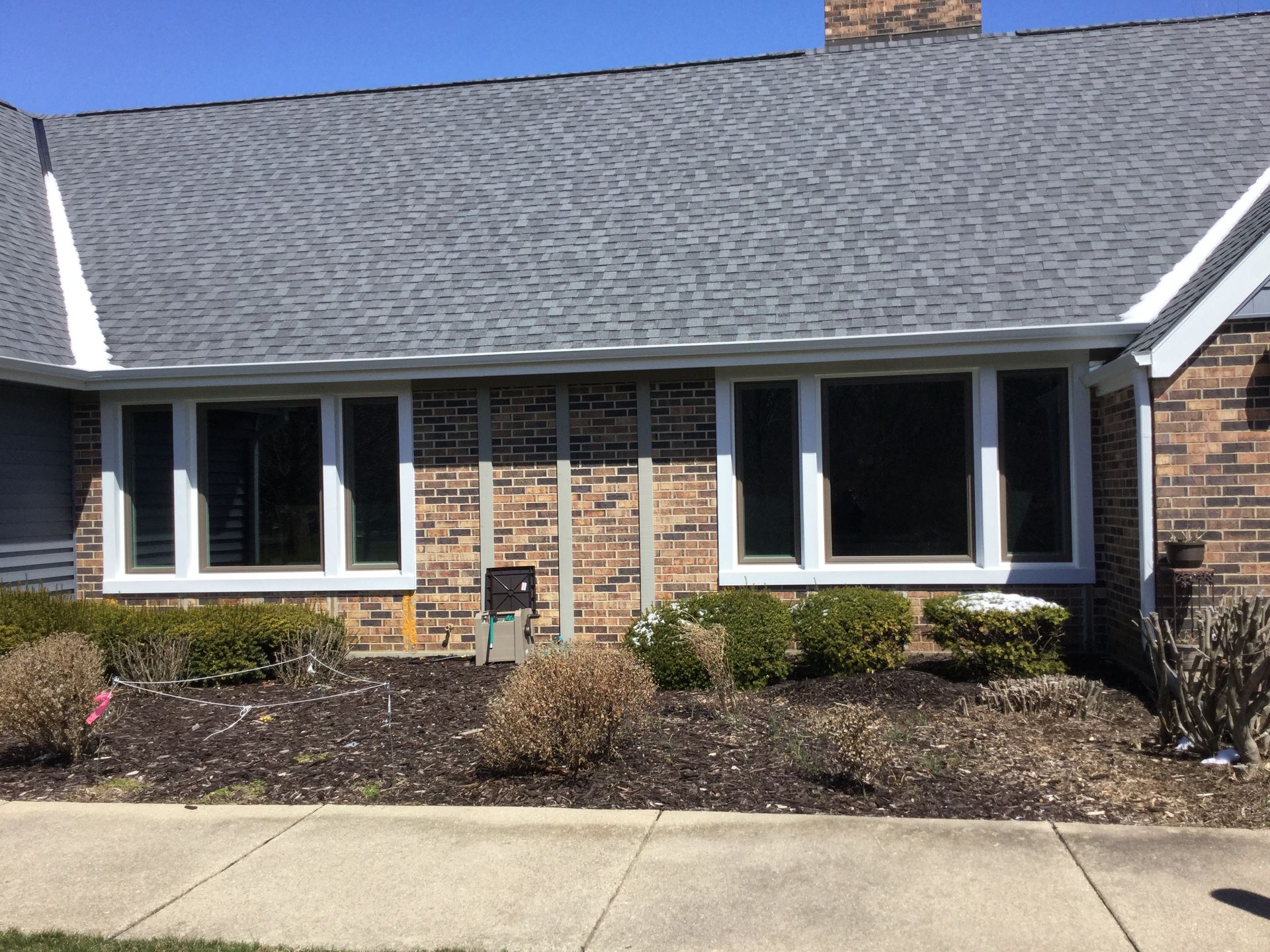 A multi-pane window set in a brick wall below a gray shingled roof, with low green shrubs in a mulch garden bed.