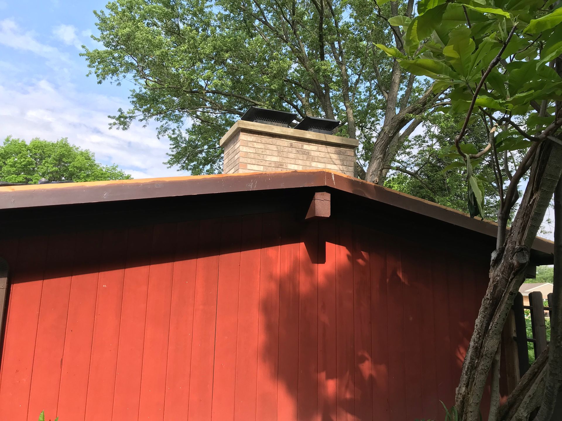 A tan stone chimney rises from the peak of a red, wood-paneled building roof, framed by green trees against a blue sky.