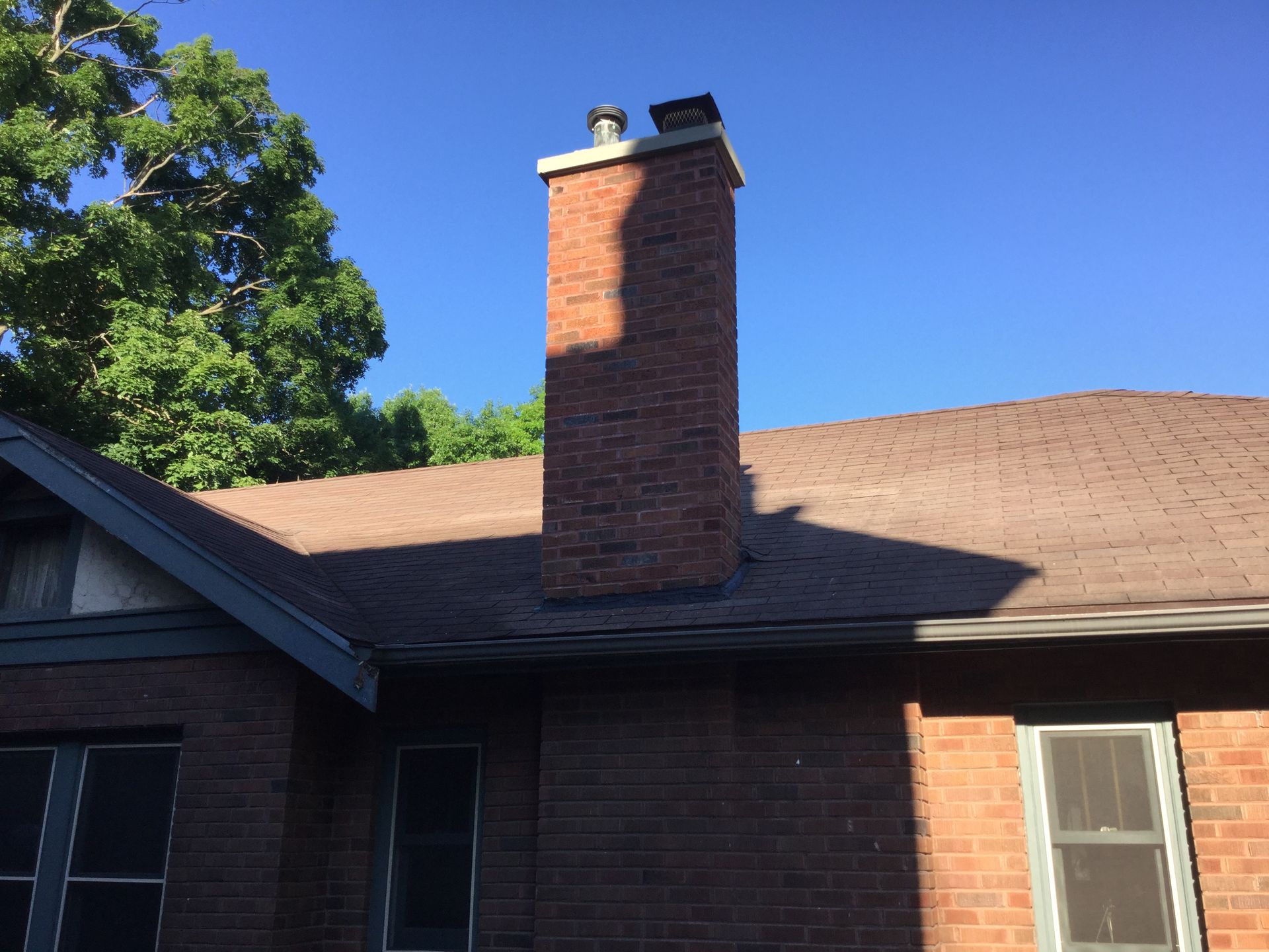 A brick chimney rises from a brown shingled roof against a clear blue sky, with trees visible in the background.