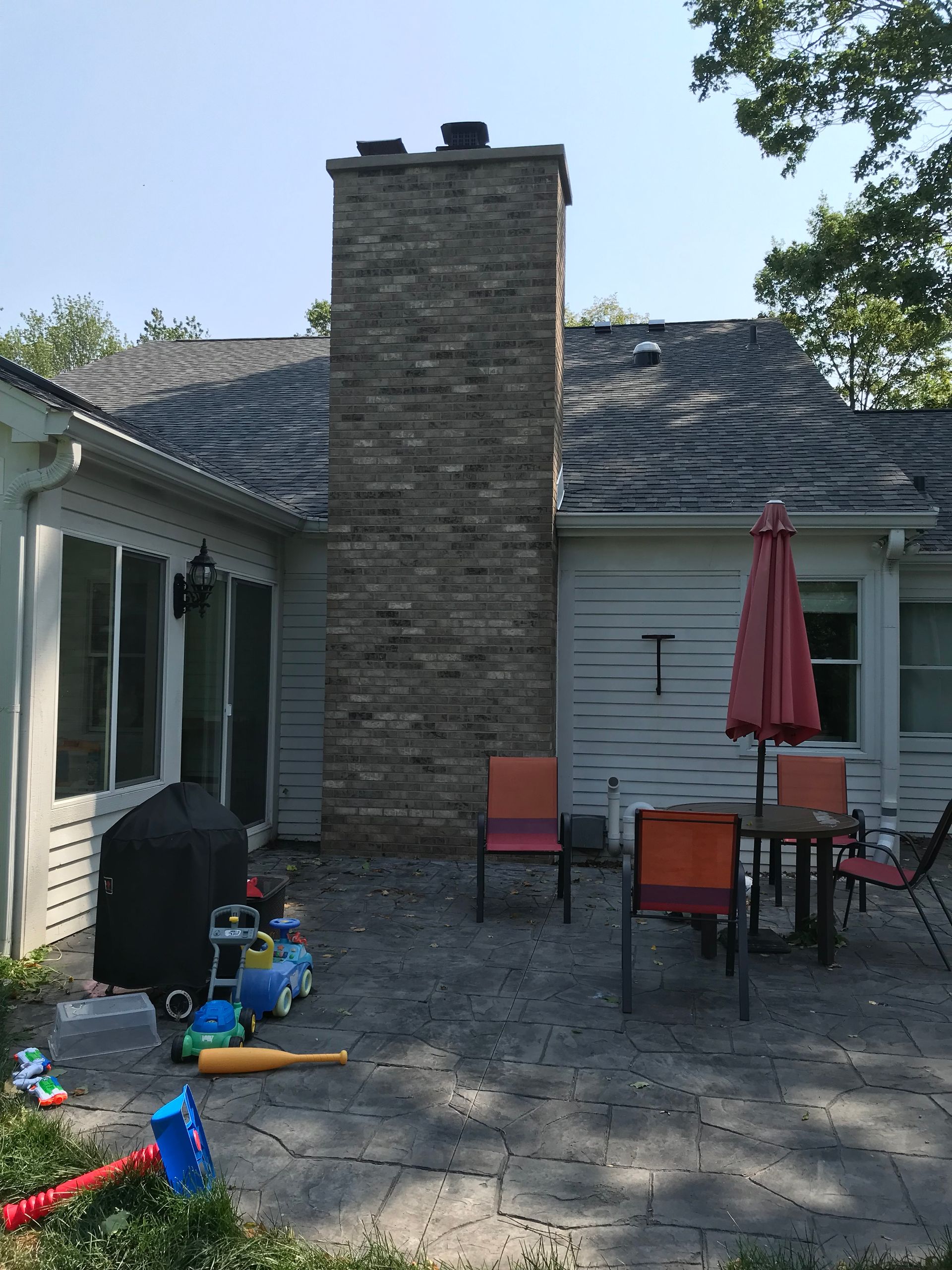 A backyard stone patio featuring a stone chimney, outdoor dining table with a red umbrella, and scattered children's toys.