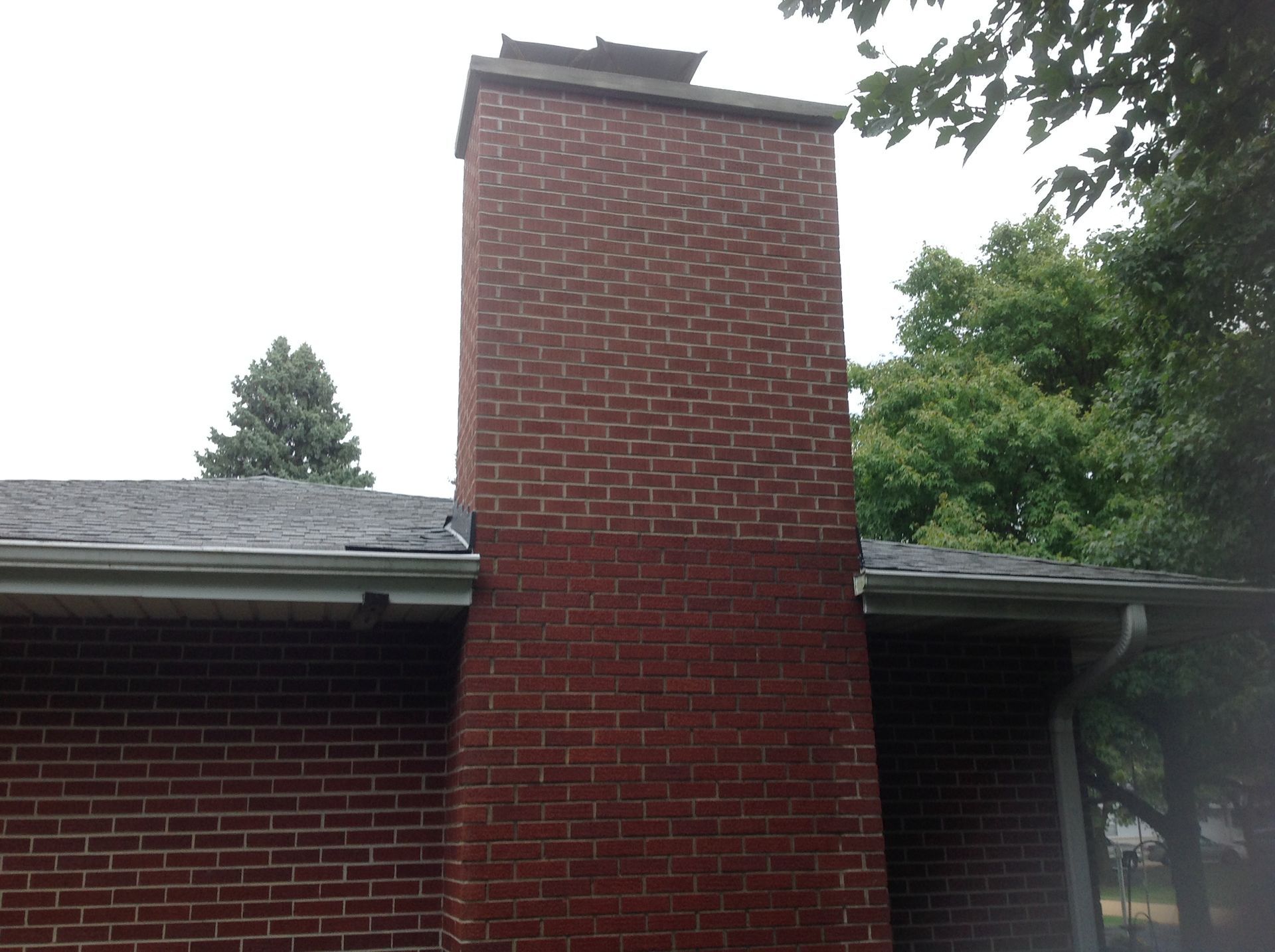 A red brick chimney rises from the gray shingled roof of a brick house under an overcast sky.