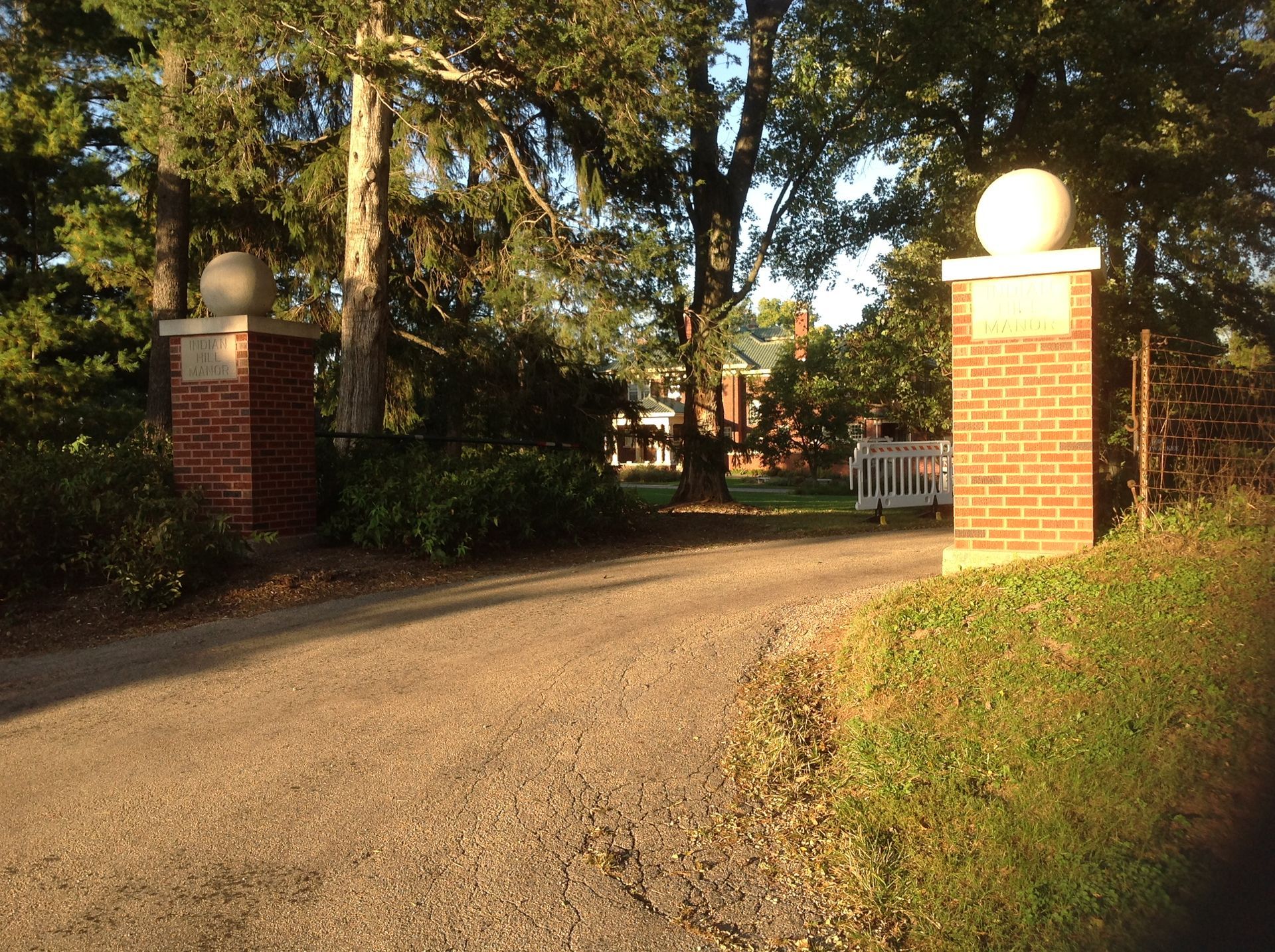A dirt driveway leads through two brick gate pillars topped with stone spheres, surrounded by mature trees and greenery.
