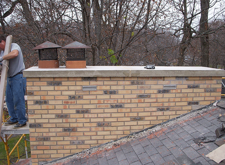 A worker stands on scaffolding next to a large brick chimney with two metal caps on a concrete crown on a rooftop.