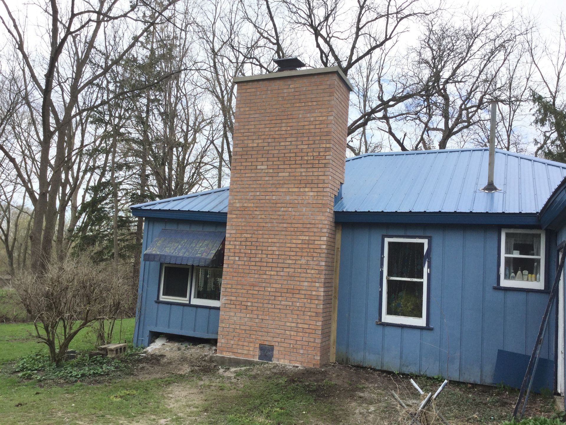 A single-story blue house with a prominent, light-colored brick chimney against a background of bare trees.