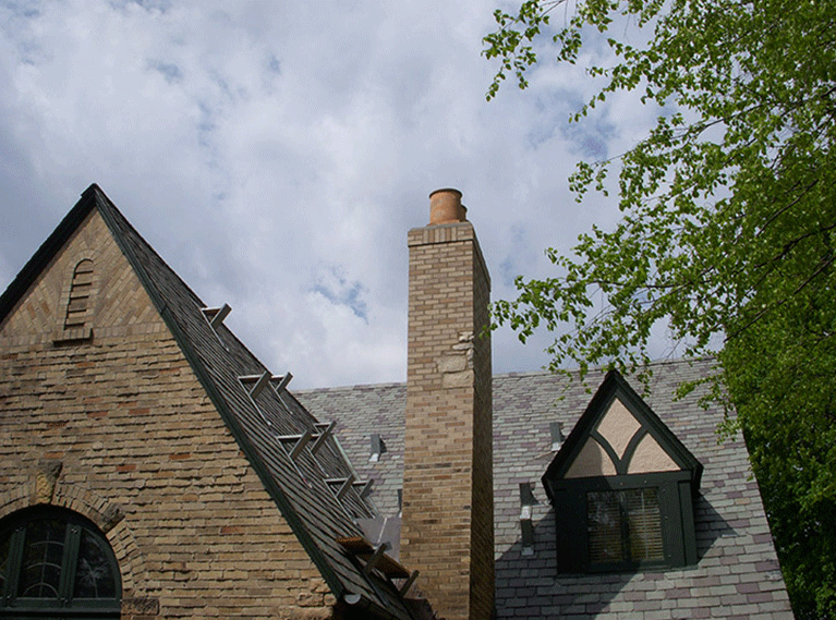 A stone cottage exterior featuring a brick chimney, steep grey roof, and a dormer window against a cloudy sky.