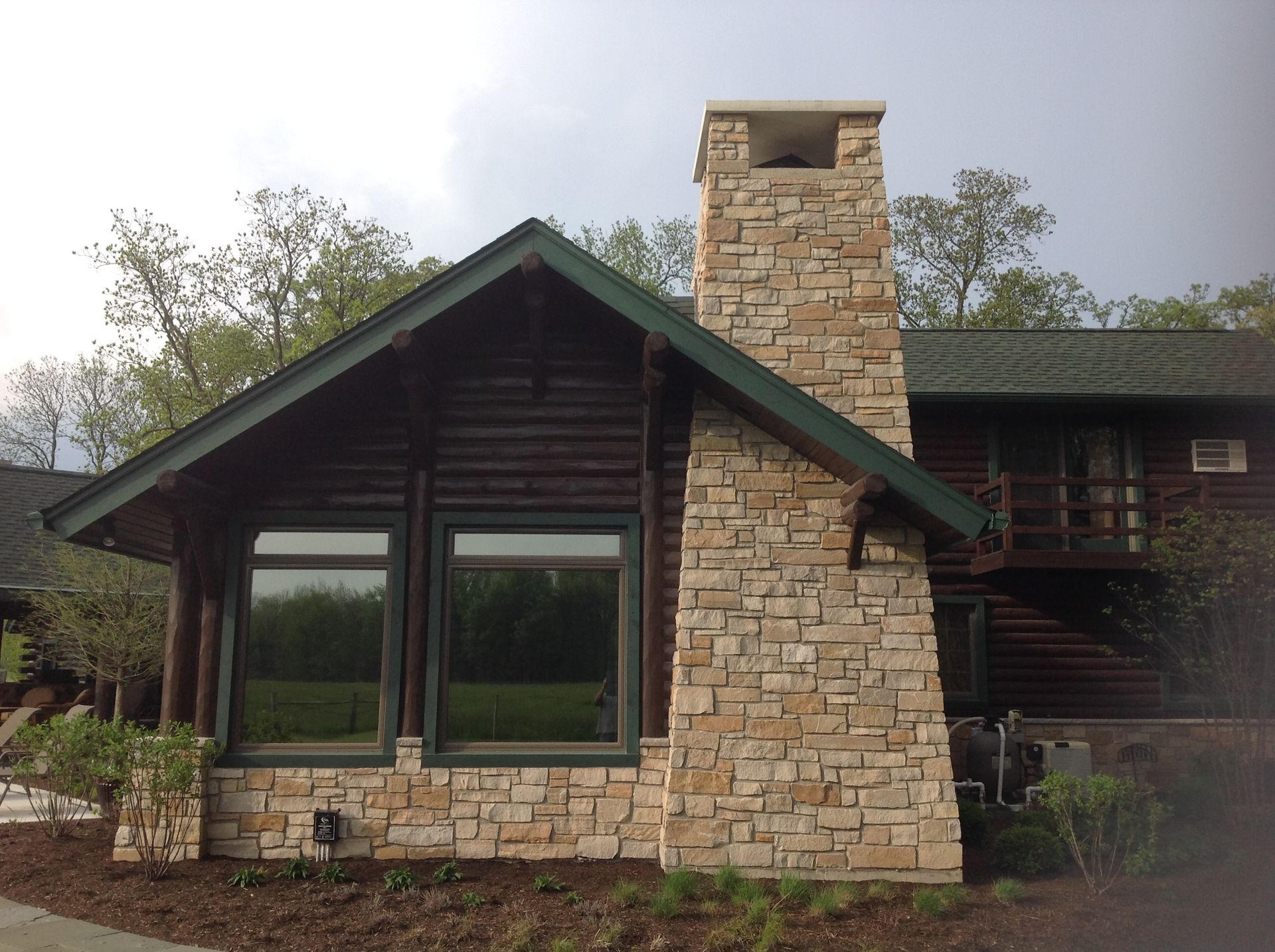 A cabin exterior featuring a large stone chimney, dark wood logs, and two windows beneath a dark green-trimmed roof.