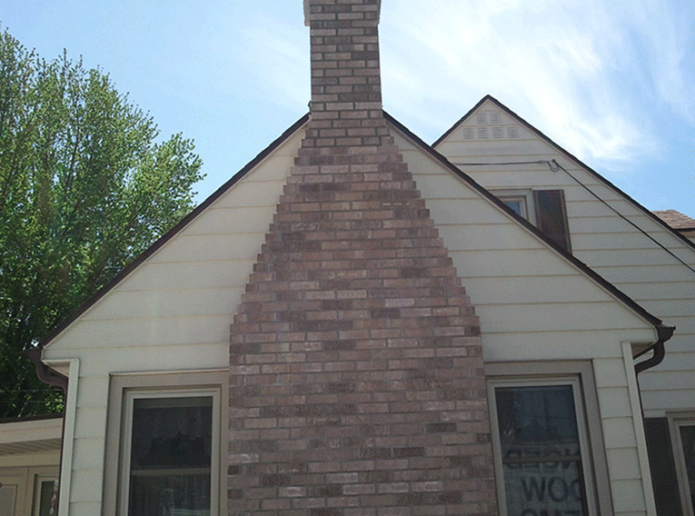 Exterior view of a house with light-colored horizontal siding and a central brick chimney tapering toward the roof peak.