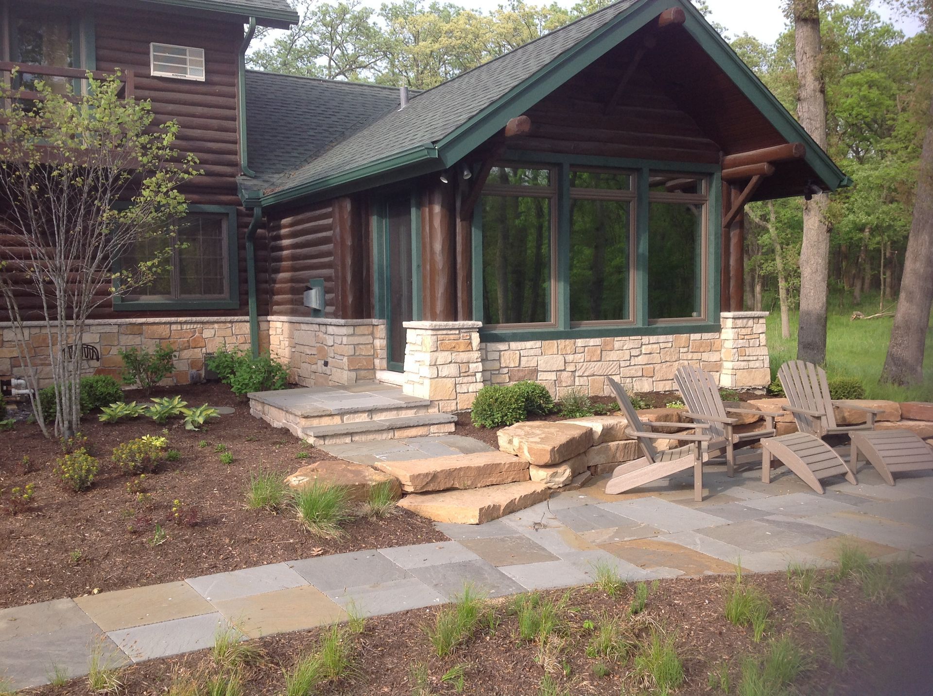 A log cabin house exterior featuring a stone patio, outdoor chairs, and a stone walkway leading to a glass-walled room.