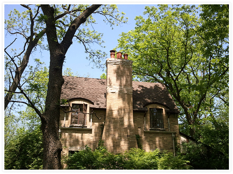 A stone cottage with a central chimney stands amidst lush green trees under a clear blue sky.