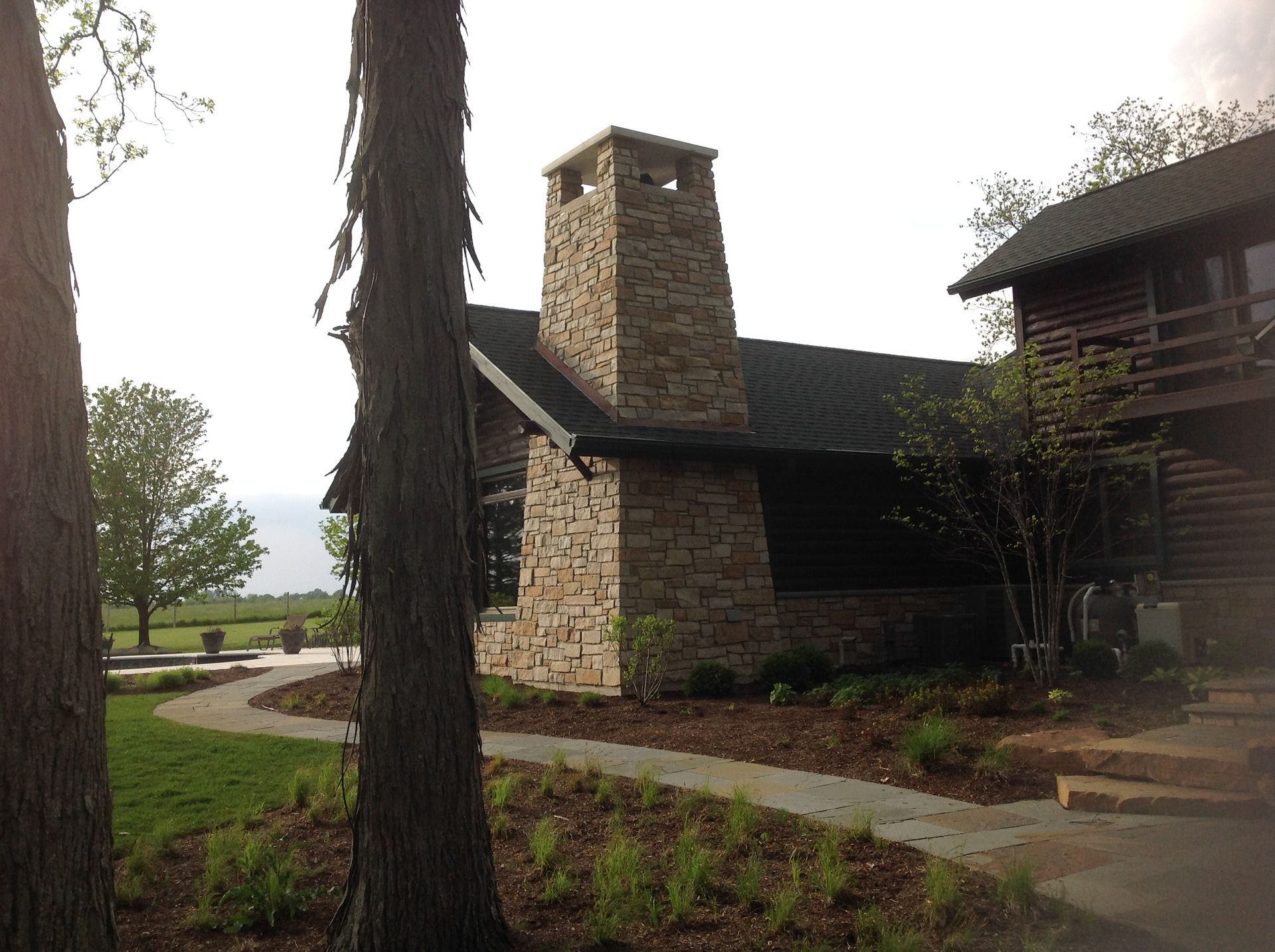 A rustic house with a large stone chimney sits in a landscaped yard, viewed past two tall tree trunks.