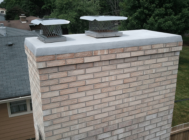 A light-colored brick chimney top featuring a gray cement crown and two metal chimney caps.