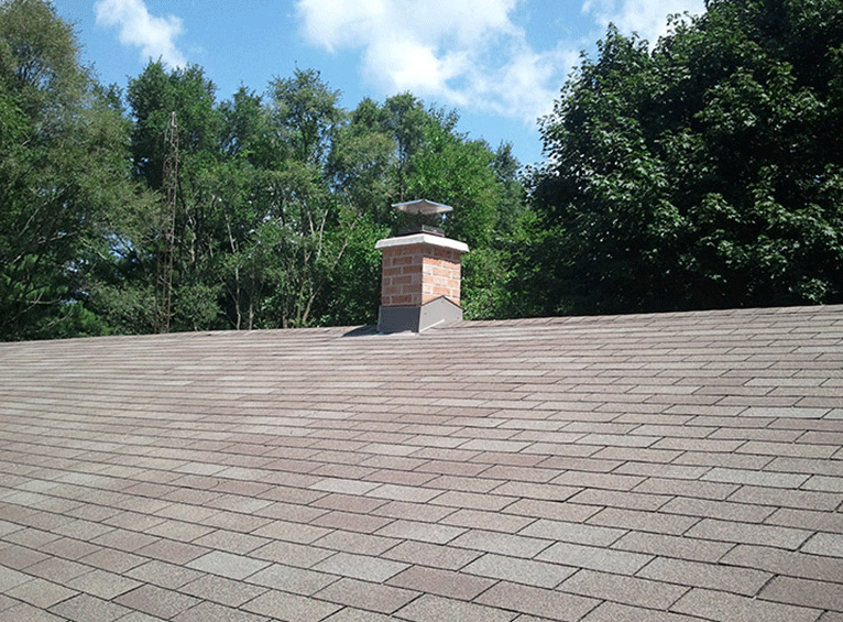 A brown asphalt shingled roof with a central brick chimney, set against a backdrop of trees and a blue sky.