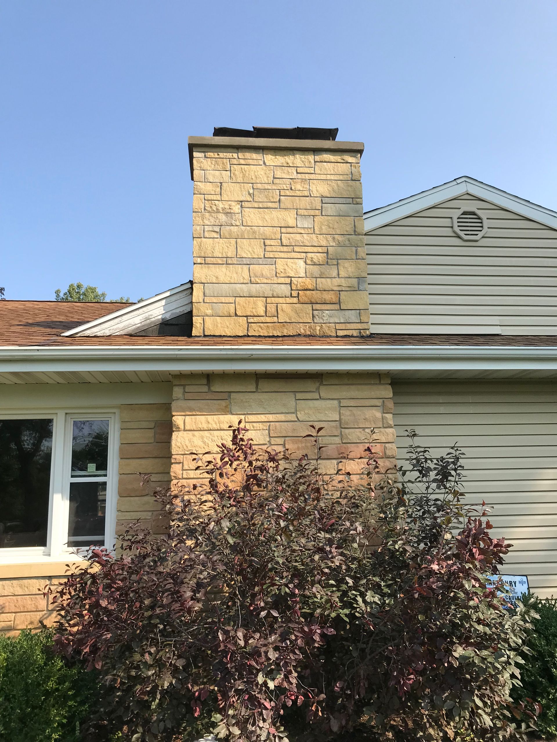 A tan stone chimney rises from the roof of a house with tan siding next to a large, dark-leafed bush.