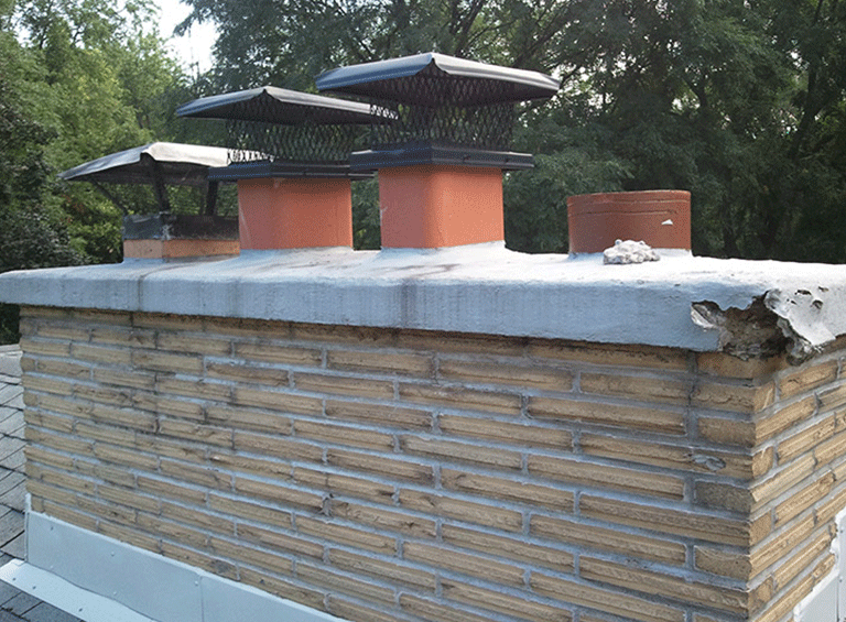 A brick chimney on a roof with four flues, three covered by metal caps and one exposed, with damaged concrete on the corner.