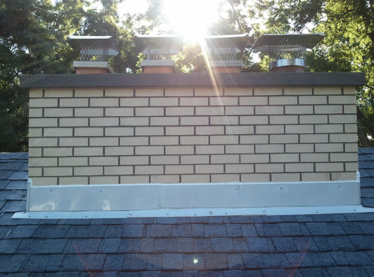 A brick chimney on a shingled roof with four metal chimney caps, seen against a sunlit sky.