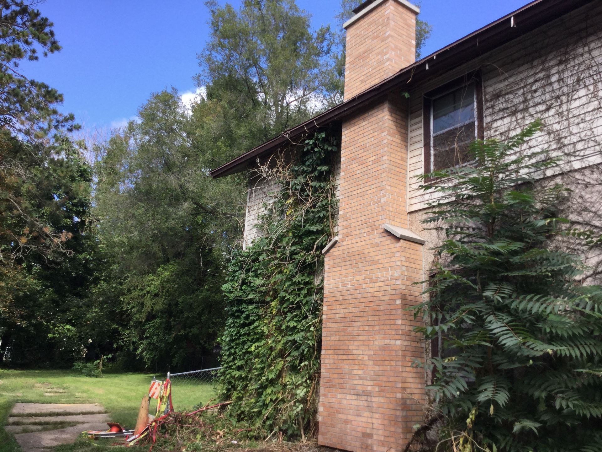 A light brown brick chimney stands against the side of a house, partially covered by climbing vines near a grassy yard.