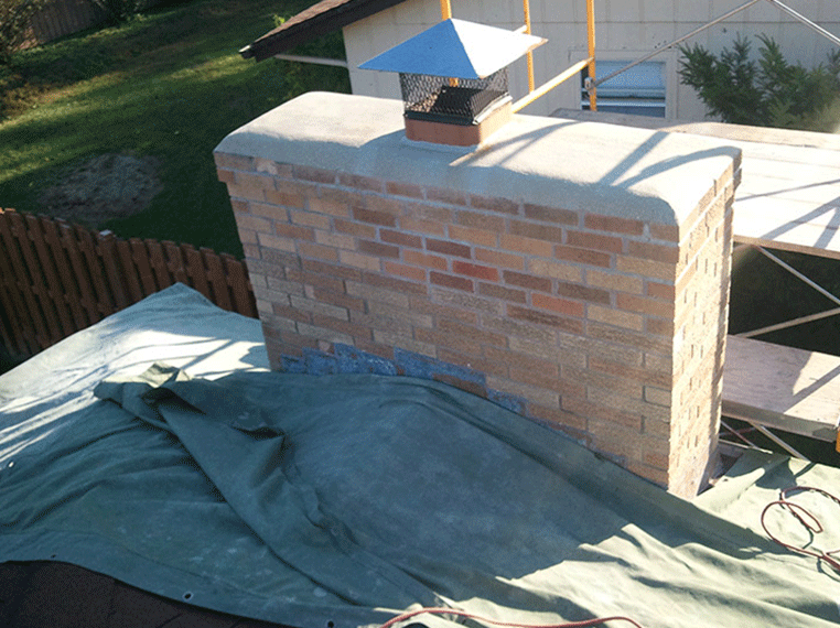 A brick chimney with a metal cap stands on a rooftop covered by a green tarp, near wooden scaffolding.