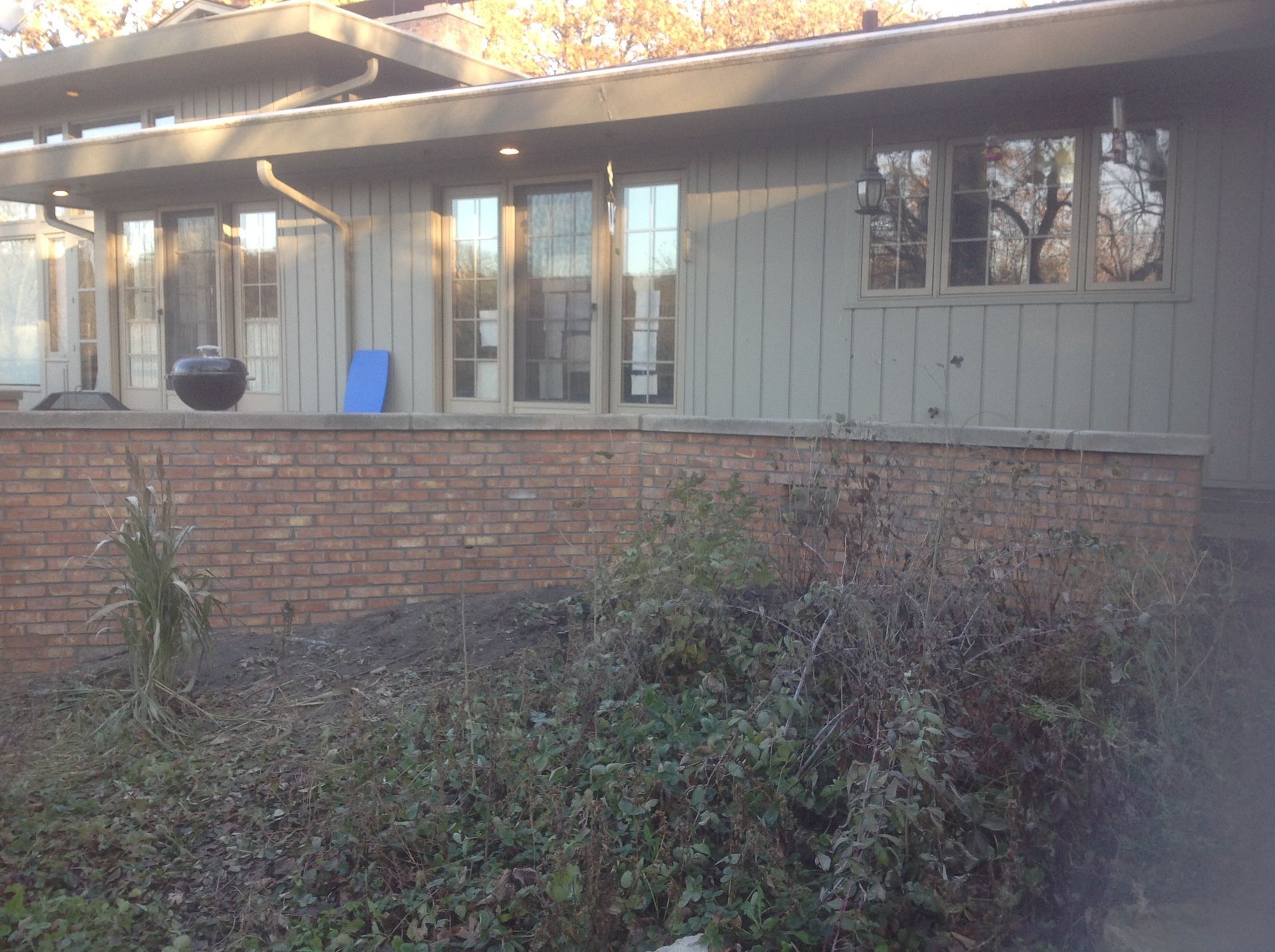 A single-story house with green vertical siding, a brick retaining wall in front, and a dark patio grill on the deck.
