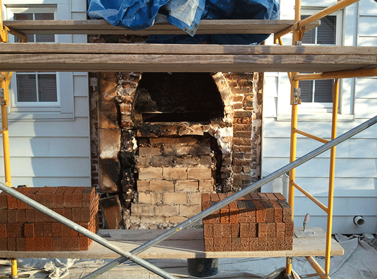 Scaffolding stands in front of a brick chimney undergoing exterior repairs, with stacks of new bricks on the platform.