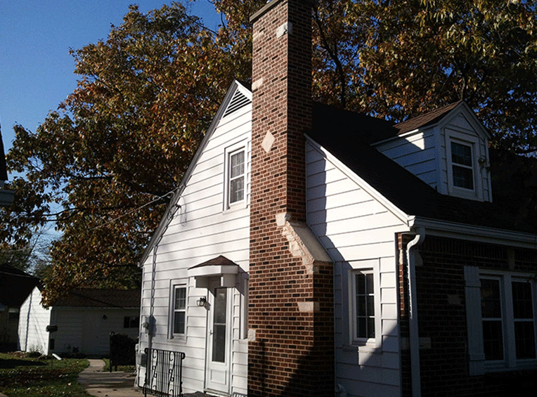A white house with a tall brick chimney and a small dormer window, set against a background of trees under a blue sky.