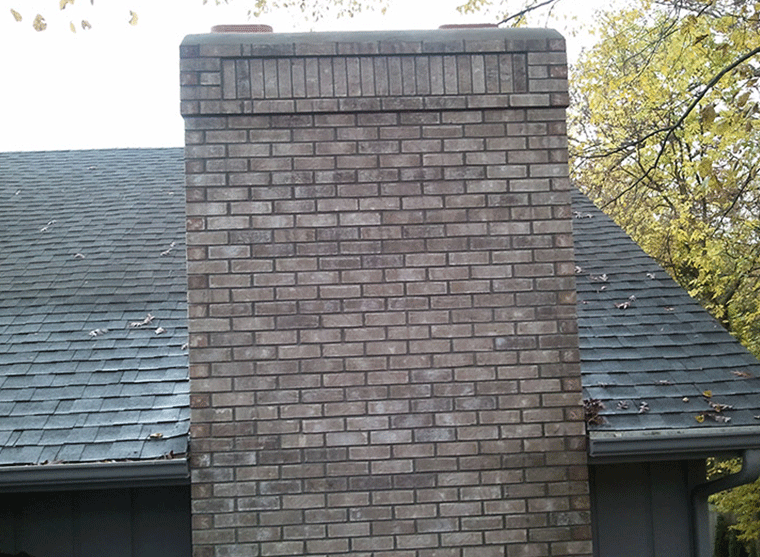 A brick chimney rises from a shingled roof against a backdrop of autumn trees.