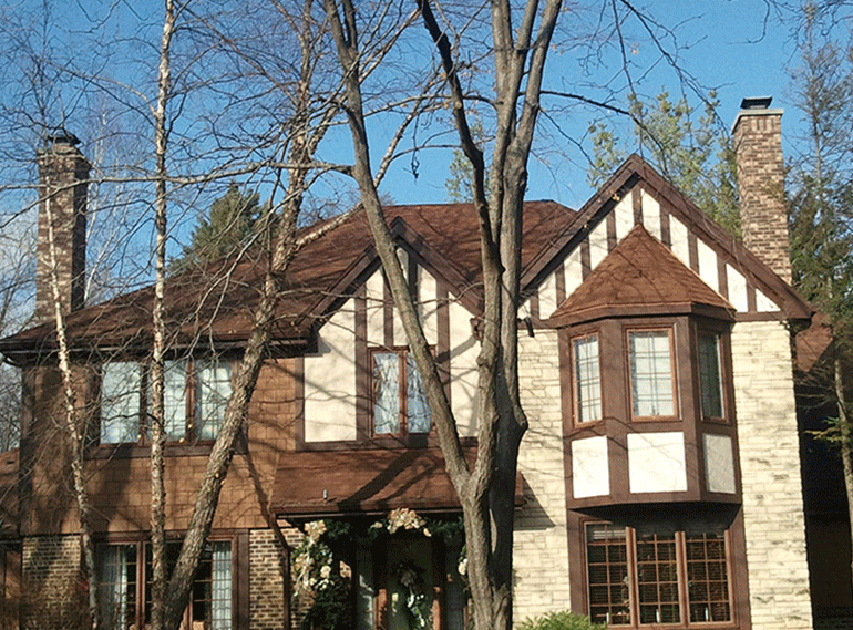 A two-story Tudor-style house with stone and brick exterior, dark wood trim, and tall chimneys, seen through bare trees.