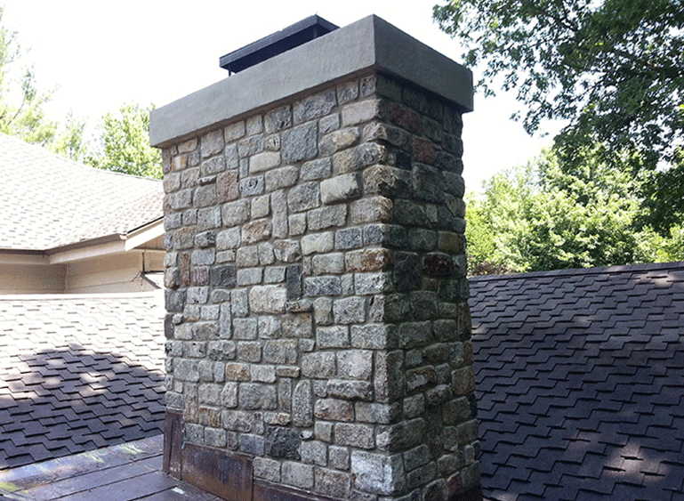 A stone chimney with a concrete cap situated on a residential roof with asphalt shingles.
