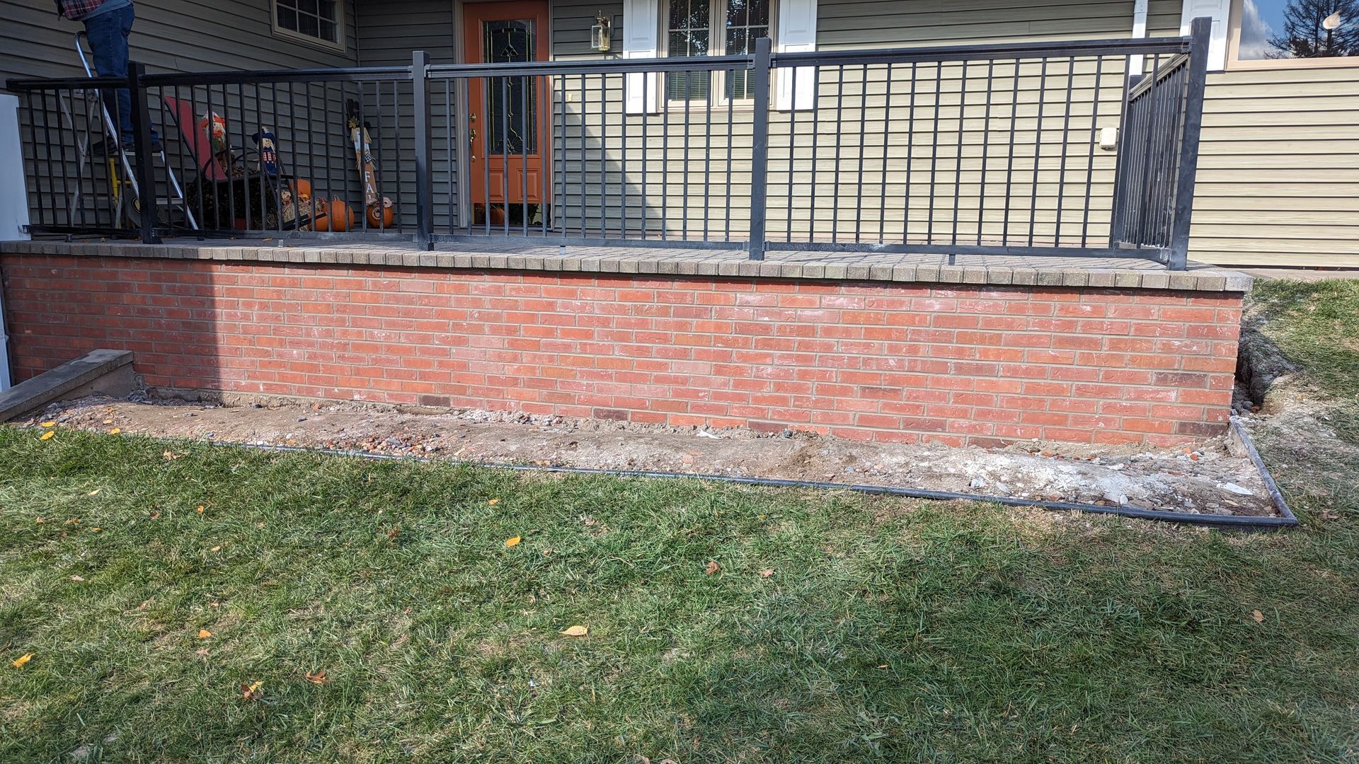 A red brick porch base with decorative black metal railing in front of a house entrance and wooden stairs.