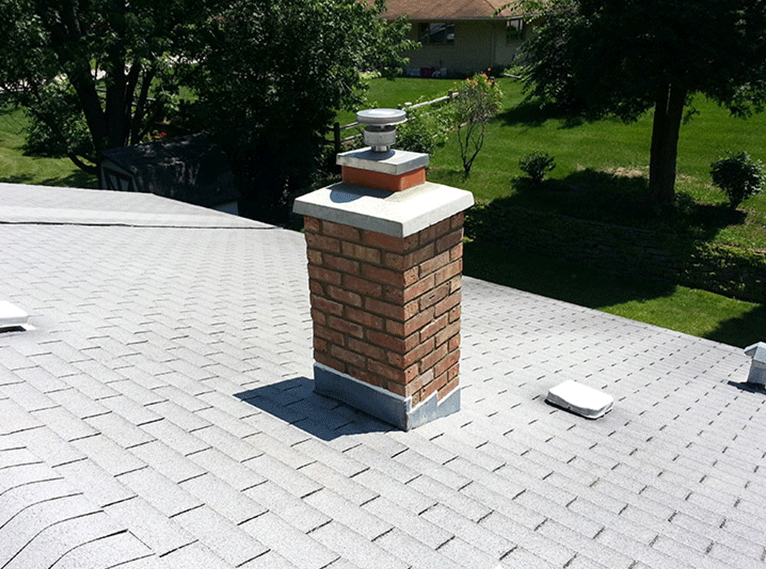 A brick chimney with a metal cap stands on a gray shingled roof under a bright, sunny sky.