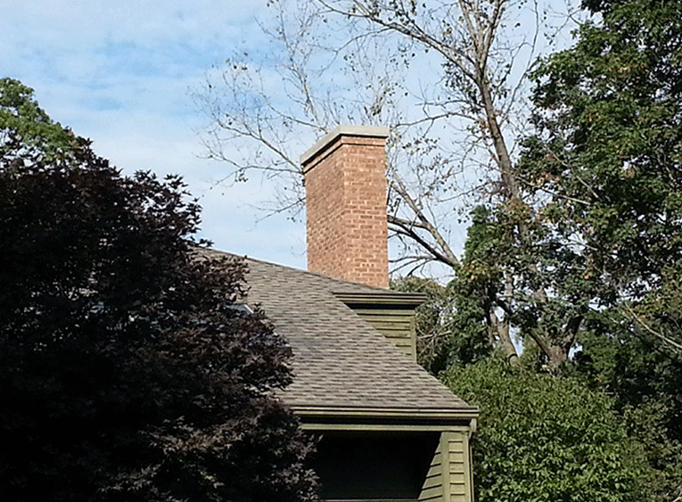 A tall brick chimney extends from a shingled roof surrounded by trees against a blue sky.