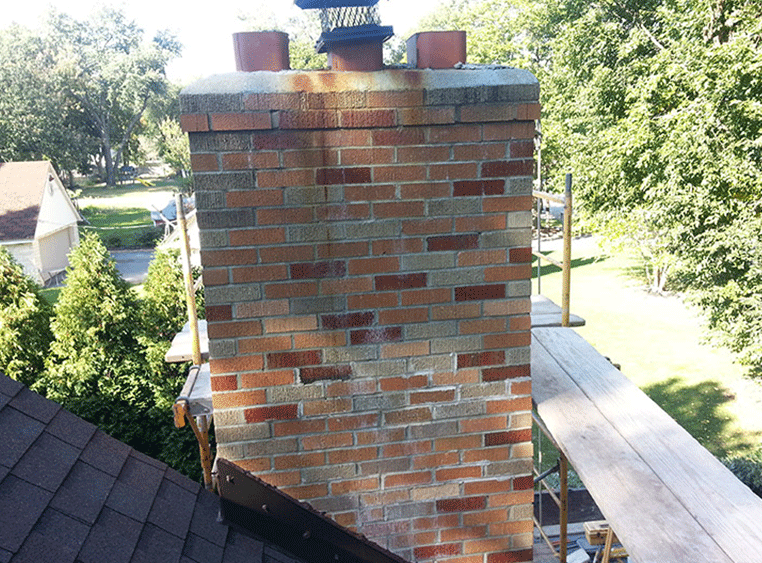 A brick chimney with rust stains near the top, surrounded by metal scaffolding on a rooftop.